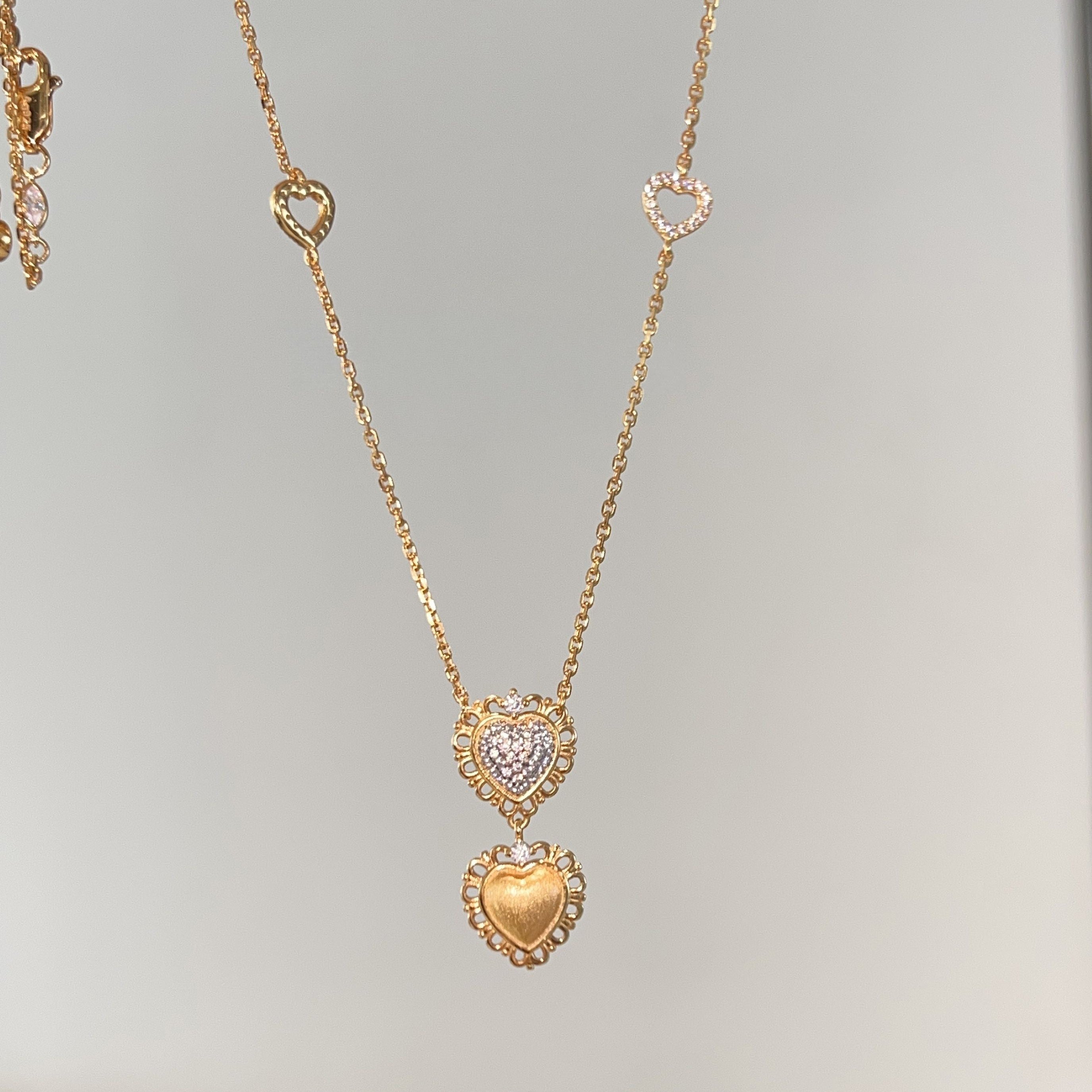 Double Heart Gold Pendant Necklace worn close-up on model — warm gold chain with cold-tone lab diamond pavé heart layered on brushed matte gold heart, showing two-tone depth and kinetic drape against collarbone
