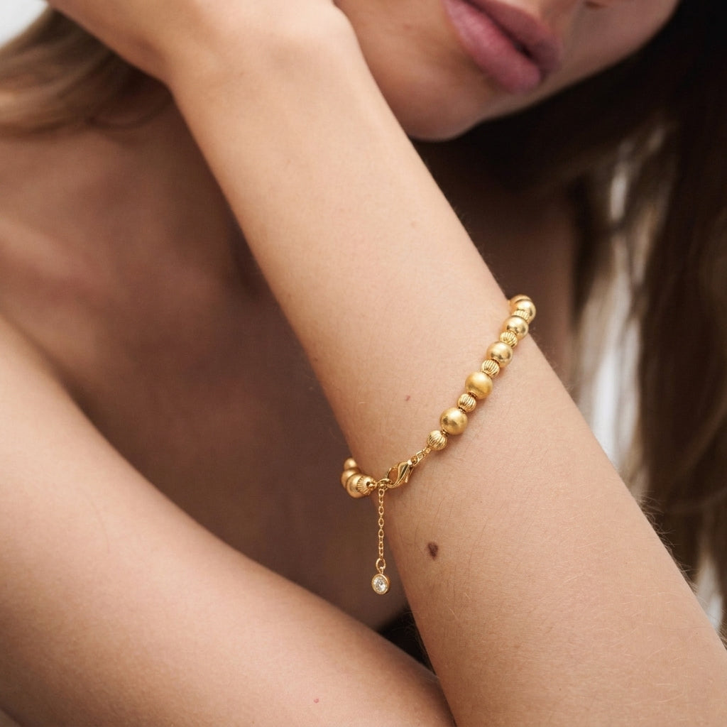 Close-up detail of gold bead bracelet — large smooth sphere beads alternating with miniature spiral-threaded spacer beads, lobster clasp with extension chain and round diamond-accented tag, matte gold finish, elastic core visible between beads