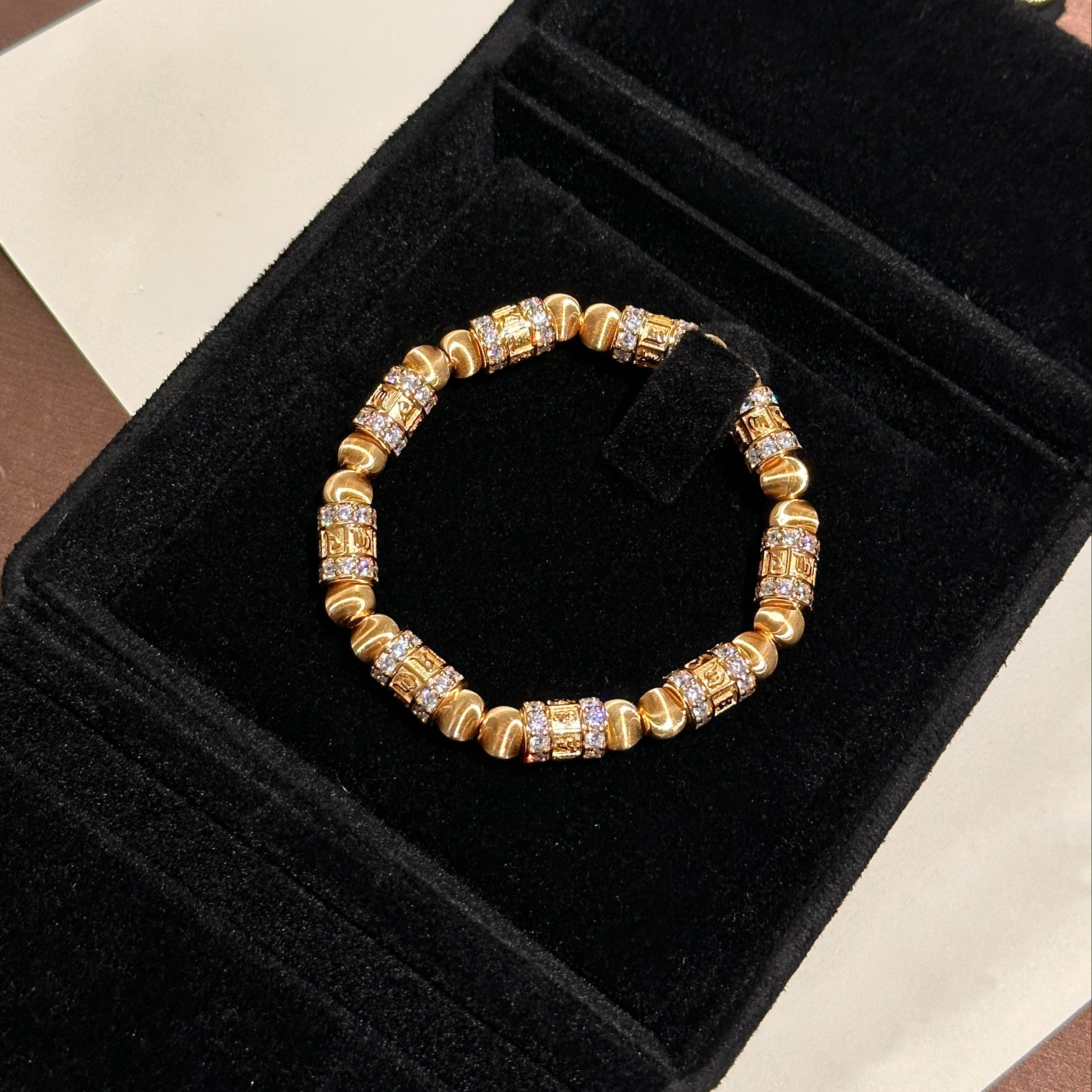 Detail shot showing single prayer wheel bead — barrel form with six-syllable mantra carved in raised relief on matte gold surface, ring of high-carbon lab diamonds in micro-claw setting at bead end, brushed gold spacer visible adjacent