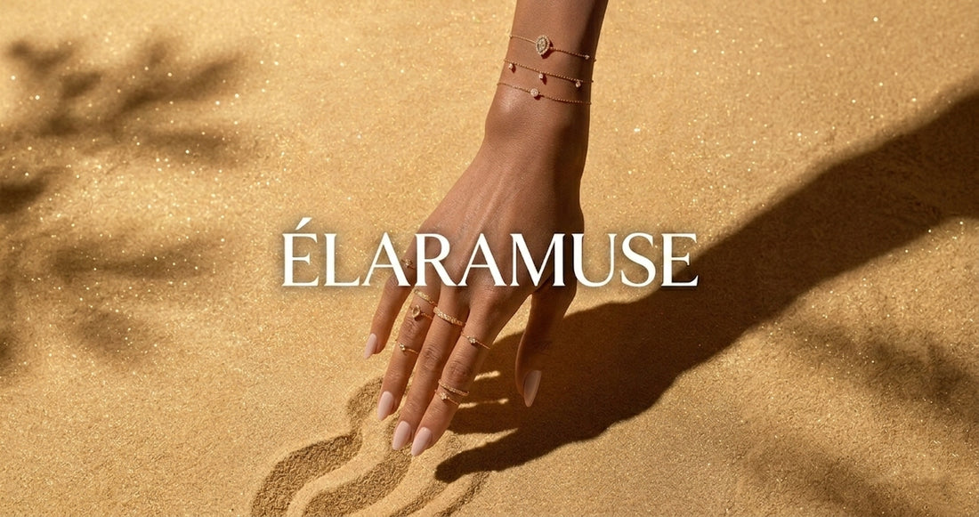 A woman's hand wearing delicate ÉLARAMUSE 18k gold rings and bracelets, gently touching golden sand. The official social sharing cover for ÉLARAMUSE everyday fine jewelry.